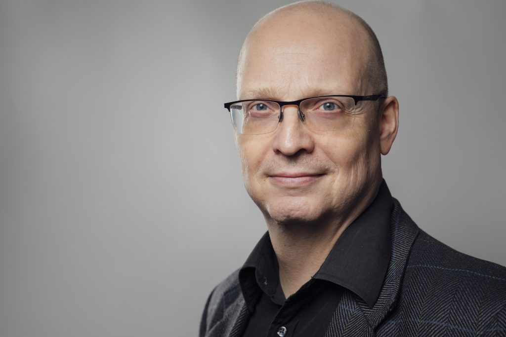 Mies tummassa paidassa harmaata taustaa vasten katsoo päättäväisin ilmein kohti kameraa.