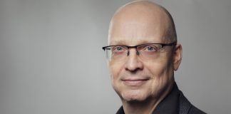 Mies tummassa paidassa harmaata taustaa vasten katsoo päättäväisin ilmein kohti kameraa.