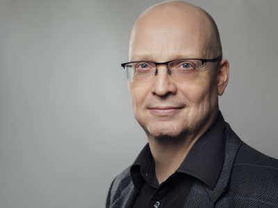 Mies tummassa paidassa harmaata taustaa vasten katsoo päättäväisin ilmein kohti kameraa.