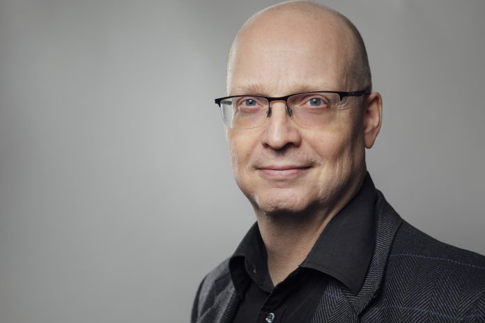 Mies tummassa paidassa harmaata taustaa vasten katsoo päättäväisin ilmein kohti kameraa.
