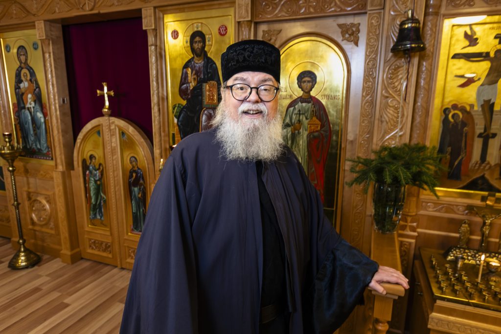 Ortodoksiseen papin asuun pukeutunut harmaapartainen mies seisoo kirkon ikonostaasin edessä ja hymyilee kameraan. Taustalla näkyy kultasävyisiä ikoneita, kynttilöitä ja puukaiverruksia.