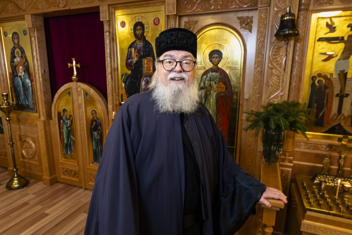 Ortodoksiseen papin asuun pukeutunut harmaapartainen mies seisoo kirkon ikonostaasin edessä ja hymyilee kameraan. Taustalla näkyy kultasävyisiä ikoneita, kynttilöitä ja puukaiverruksia.