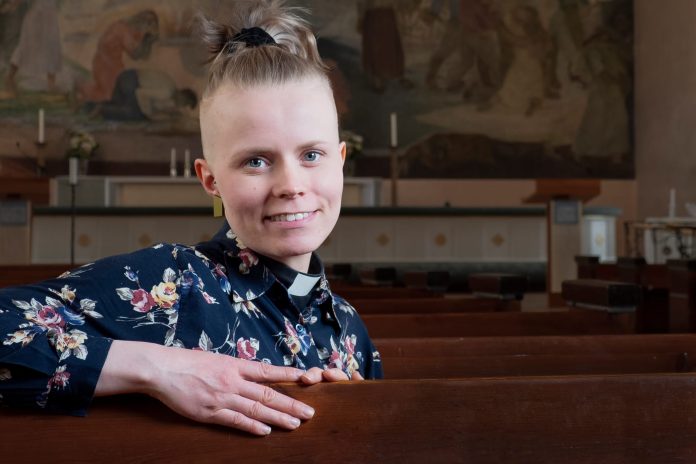 Kuvassa nuori nainen istuu kirkon penkissä ja katsoo suoraan kameraan.