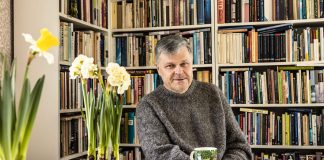 Kolumni: Keskustelu tekoäly-Jeesuksen kanssa oli hämmentävä kokemus Kuvassa mies istuu pöydän ääressä, takana on kirjahylly täynnä kirjoja. Pöydällä miehen edessä on vihreäsävyinen muumimuki ja narssisseja.
