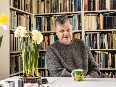 Kuvassa mies istuu pöydän ääressä, takana on kirjahylly täynnä kirjoja. Pöydällä miehen edessä on vihreäsävyinen muumimuki ja narssisseja.