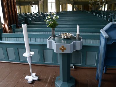 Vaaleansinisen puukirkon sisätila nähtynä alttarin suunnasta. Etualalla on kastemalja, jonka päällä on kukkakimppu, kynttilä ja metallinen malja. Taustalla näkyy pitkiä kirkonpenkkirivejä ja korkeat ikkunat, joista tulee luonnonvaloa.