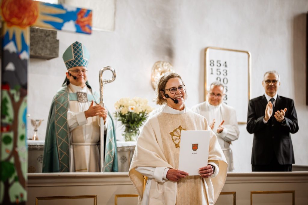 Papiksi pukeutunut henkilö seisoo kirkon etuosassa ja pitelee asiakirjaa. Taustalla alttarilla seisoo piispan asuun pukeutunut henkilö sauvoineen sekä kaksi muuta henkilöä, jotka taputtavat. Kirkon sisätila on valoisa, ja etualalla näkyy osittain värikäs koriste.