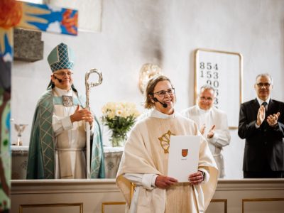 Papiksi pukeutunut henkilö seisoo kirkon etuosassa ja pitelee asiakirjaa. Taustalla alttarilla seisoo piispan asuun pukeutunut henkilö sauvoineen sekä kaksi muuta henkilöä, jotka taputtavat. Kirkon sisätila on valoisa, ja etualalla näkyy osittain värikäs koriste.