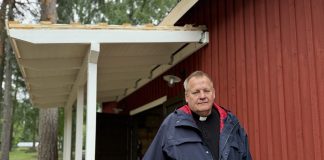 Pappi seisoo punaisen puurakennuksen kuistilla ja nojaa puiseen kaiteeseen.