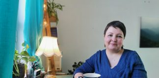 Henkilö istuu siniseksi maalatun pöydän ääressä ja pitää käsissään kuvioitua mukia. Ikkunasta tuleva valo, turkoosit verhot, viherkasvit ja pöytälamppu luovat kodikkaan tunnelman.