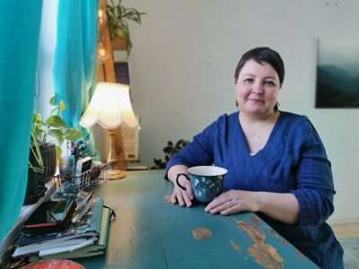 Henkilö istuu siniseksi maalatun pöydän ääressä ja pitää käsissään kuvioitua mukia. Ikkunasta tuleva valo, turkoosit verhot, viherkasvit ja pöytälamppu luovat kodikkaan tunnelman.