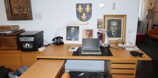 Kuvassa on presidentti Urho Kekkosen työpöytä, jonka taustalla on useita valokuvia Kekkosesta sekä hänen itse puutauluun tekemä tyylitelty omakuva itsestään. Ruskealla pöydällä on vanhanmallinen musta lankapuhelin. Seinällä on vaakuna, jossa on kolme kultaista kuusta.
