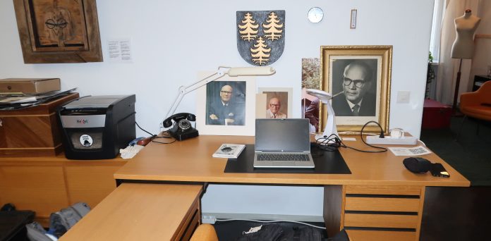 Kuvassa on presidentti Urho Kekkosen työpöytä, jonka taustalla on useita valokuvia Kekkosesta sekä hänen itse puutauluun tekemä tyylitelty omakuva itsestään. Ruskealla pöydällä on vanhanmallinen musta lankapuhelin. Seinällä on vaakuna, jossa on kolme kultaista kuusta.