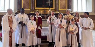 Ryhmä lapsia, nuoria ja yksi aikuinen seisoo kirkon alttarin edessä liturgisissa asuissa. Osa pitää käsissään kynttilöitä ja kulkuristejä. Taustalla näkyy alttari sekä suuri uskonnollinen maalaus.