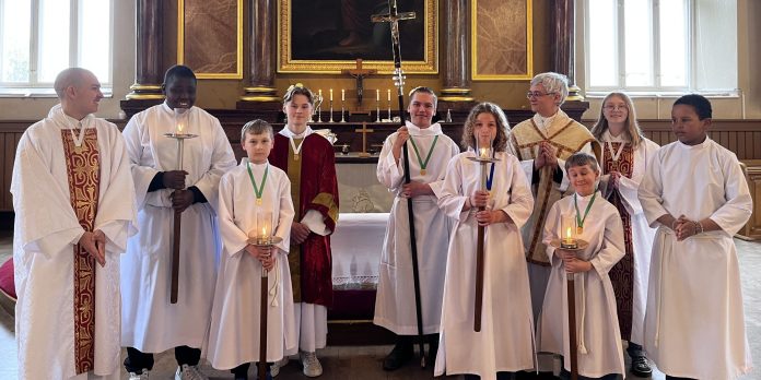 Ryhmä lapsia, nuoria ja yksi aikuinen seisoo kirkon alttarin edessä liturgisissa asuissa. Osa pitää käsissään kynttilöitä ja kulkuristejä. Taustalla näkyy alttari sekä suuri uskonnollinen maalaus.