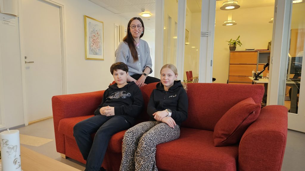 Kolme henkilöä toimistomaisessa sisätilassa. Kaksi nuorta istuu punaisella sohvalla ja heidän takanaan seisoo aikuinen henkilö. Taustalla näkyy käytävä, toimistohuoneita lasiseinineen, valaisimia sekä seinällä kehystetty taulu.