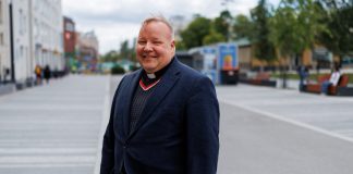 Pappi seisoo kaupunkiaukiolla ja hymyilee kameraan. Hänellä on yllään tumma takki ja papinpaita valkoisella papinkauluksella. Taustalla näkyy kävelyalue, rakennuksia, puita ja muutamia ihmisiä.