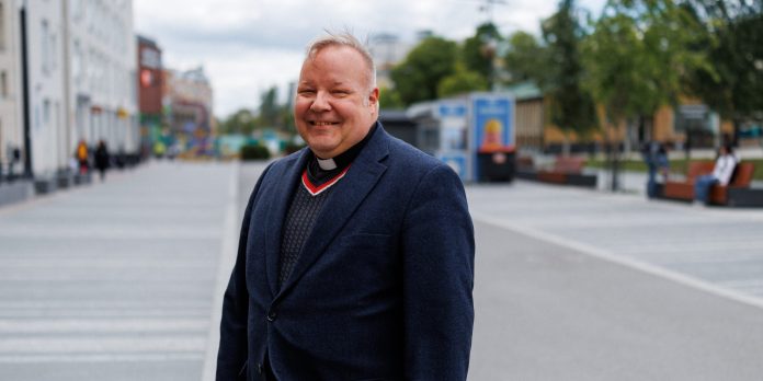 Pappi seisoo kaupunkiaukiolla ja hymyilee kameraan. Hänellä on yllään tumma takki ja papinpaita valkoisella papinkauluksella. Taustalla näkyy kävelyalue, rakennuksia, puita ja muutamia ihmisiä.