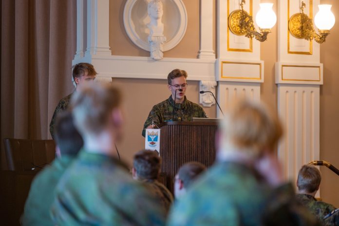 Maastopukuinen Arttu Vuori puhuu puhujakorokkeella salissa, jossa maastopukuihin sonnustautunut yleisö kuuntelee istuen.
