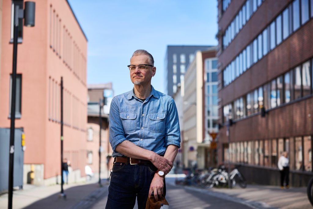 silmälasipäinen mies seisoo kadulla kahden rakennuksen välissä ja katsoo kameraan