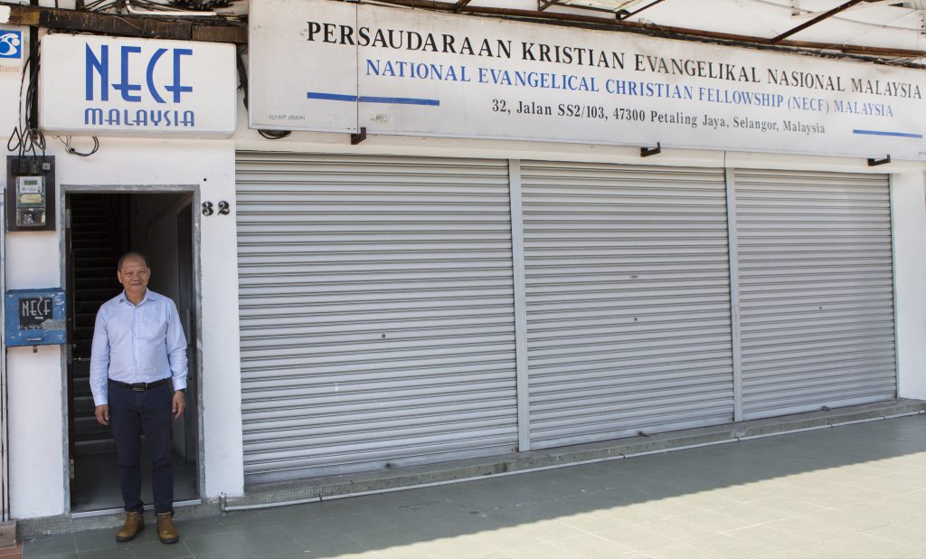 Henkilö seisoo avoimessa oviaukossa rakennuksen edessä, jonka yläpuolella on National Evangelical Christian Fellowship (NECF) Malaysia -järjestön kyltti. Rakennuksen metalliset rullaovet ovat suljettuina, ja sisäänkäynnin vieressä näkyy talonumero 32.