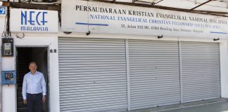 Henkilö seisoo avoimessa oviaukossa rakennuksen edessä, jonka yläpuolella on National Evangelical Christian Fellowship (NECF) Malaysia -järjestön kyltti. Rakennuksen metalliset rullaovet ovat suljettuina, ja sisäänkäynnin vieressä näkyy talonumero 32.