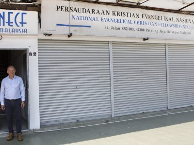 Henkilö seisoo avoimessa oviaukossa rakennuksen edessä, jonka yläpuolella on National Evangelical Christian Fellowship (NECF) Malaysia -järjestön kyltti. Rakennuksen metalliset rullaovet ovat suljettuina, ja sisäänkäynnin vieressä näkyy talonumero 32.