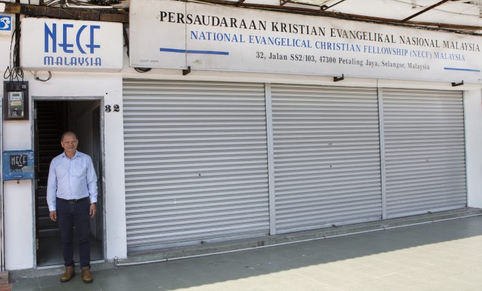 Henkilö seisoo avoimessa oviaukossa rakennuksen edessä, jonka yläpuolella on National Evangelical Christian Fellowship (NECF) Malaysia -järjestön kyltti. Rakennuksen metalliset rullaovet ovat suljettuina, ja sisäänkäynnin vieressä näkyy talonumero 32.