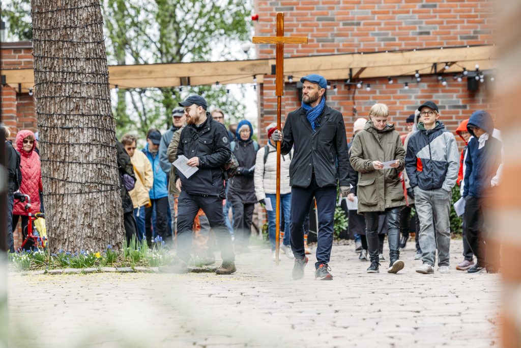 Joukko ihmisiä kävelee pihatiellä ulkotapahtumassa. Etualalla henkilö kantaa suurta puista ristiä, ja osallistujilla on käsissään paperiarkkeja. Taustalla näkyy tiilirakennus, puu ja ulkovalaisimia.
