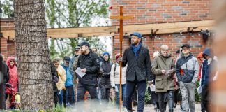 Joukko ihmisiä kävelee pihatiellä ulkotapahtumassa. Etualalla henkilö kantaa suurta puista ristiä, ja osallistujilla on käsissään paperiarkkeja. Taustalla näkyy tiilirakennus, puu ja ulkovalaisimia.