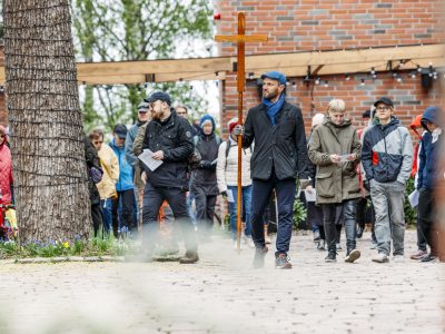 Joukko ihmisiä kävelee pihatiellä ulkotapahtumassa. Etualalla henkilö kantaa suurta puista ristiä, ja osallistujilla on käsissään paperiarkkeja. Taustalla näkyy tiilirakennus, puu ja ulkovalaisimia.