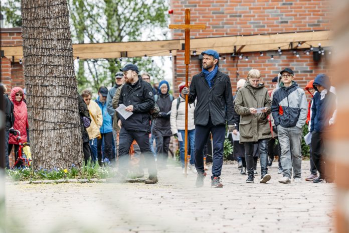 Joukko ihmisiä kävelee pihatiellä ulkotapahtumassa. Etualalla henkilö kantaa suurta puista ristiä, ja osallistujilla on käsissään paperiarkkeja. Taustalla näkyy tiilirakennus, puu ja ulkovalaisimia.