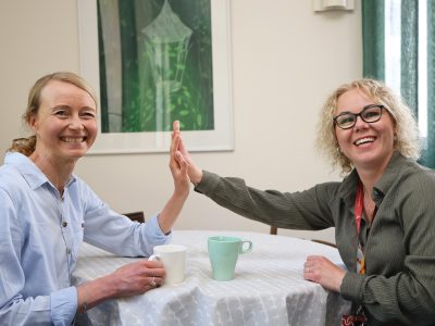 Kaksi hymyilevää naista istuu vastakkain pyöreän pöydän ääressä ja antaa toisilleen ylävitosen. Pöydällä on kaksi kahvimukia, ja taustalla näkyy kehystetty vihreäsävyinen taulu sekä verhot.