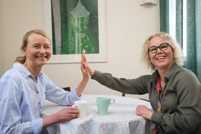 Kaksi hymyilevää naista istuu vastakkain pyöreän pöydän ääressä ja antaa toisilleen ylävitosen. Pöydällä on kaksi kahvimukia, ja taustalla näkyy kehystetty vihreäsävyinen taulu sekä verhot.