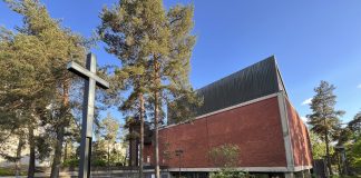 Pihlajamäen kirkko myytiin Suomen etiopialaisortodoksien yhteisölle Punatiilinen kirkkorakennus kohoaa mäntyjen keskellä aurinkoisena päivänä. Etualalla seisoo suuri musta metallinen risti, ja rakennuksen yläosa on tummaa pystylaudoitusta. Kirkasta sinistä taivasta vasten näkyvät korkeat puut kehystävät rakennusta.