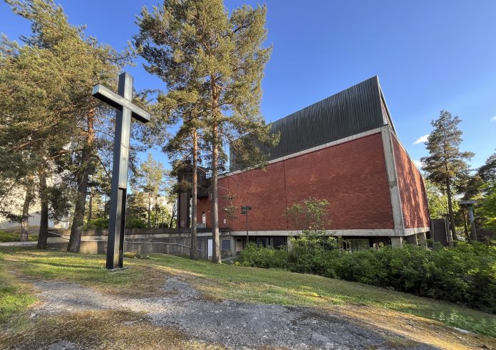 Punatiilinen kirkkorakennus kohoaa mäntyjen keskellä aurinkoisena päivänä. Etualalla seisoo suuri musta metallinen risti, ja rakennuksen yläosa on tummaa pystylaudoitusta. Kirkasta sinistä taivasta vasten näkyvät korkeat puut kehystävät rakennusta.