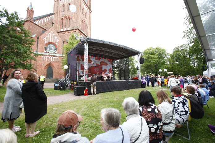 Vanhempia naisia istuu katsomossa lavan edessä. Taustalla näkyy Keski-Porin kirkko.