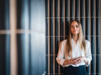 Vaaleaan puseroon pukeutunut pitkähiuksinen henkilö seisoo sisätilassa tumman pystylaatoitetun seinän edessä ja katsoo kameraan.