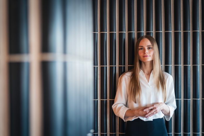 Vaaleaan puseroon pukeutunut pitkähiuksinen henkilö seisoo sisätilassa tumman pystylaatoitetun seinän edessä ja katsoo kameraan.
