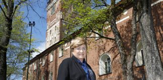 Henkilö seisoo nurmikolla punatiilisen kirkon vieressä aurinkoisena kevätpäivänä. Taustalla näkyvät kirkon korkea torni, kaarevat ikkunat ja lehtevät puut, joiden oksat kehystävät näkymää.