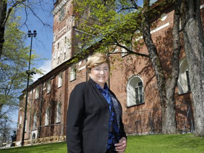 Henkilö seisoo nurmikolla punatiilisen kirkon vieressä aurinkoisena kevätpäivänä. Taustalla näkyvät kirkon korkea torni, kaarevat ikkunat ja lehtevät puut, joiden oksat kehystävät näkymää.