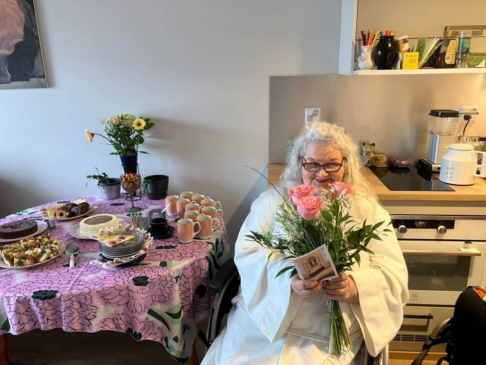 70-vuotias konfirmoitu Eija Seppänen istuu alba yllään pyörätuolissa kotonaan.