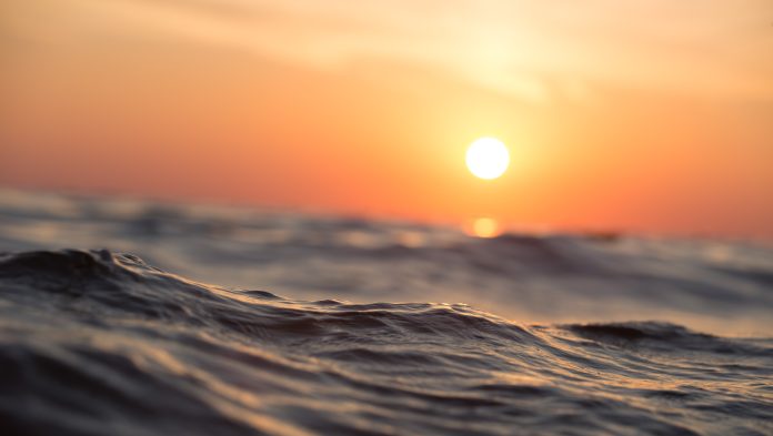 Kuvassa on aaltoilevaa merka ja aurinko ja oranssi taivas.