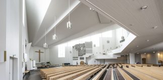Kaksi Aalto-kirkkoa jäi vaille maailmanperintökohteen asemaa – selvitimme miksi Valoisa, moderni kirkkosali, jossa vaaleat puupenkit suuntautuvat alttaria kohti. Alttarin taustalla on suuri puinen risti, ja oikealla näkyvät urut sekä korkea, viisto katto suurine ikkunoineen.