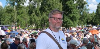 Valkoiseen T-paitaan pukeutunut pastori Simo Korkalainen Ylivieskan juhlakentällä, taustallaan paljon penkeissä istuvaa juhlakansaa.