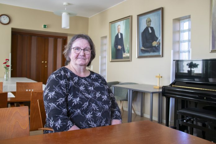 Silmälasipäinen henkilö istuu pöydän ääressä vaaleassa seurakuntasalissa ja katsoo kameraan. Taustalla näkyy musta piano, pieni puinen risti sekä seinällä kaksi kehystettyä muotokuvaa. Vasemmalla on pöytiä, tuoleja ja puiset pariovet.