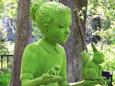 Kuvassa kauttaaltaan virhreä tyttöpatsas pitää kädellään sammakkoa ja oravaa. Taustalla on vihreää lehtimetsää.