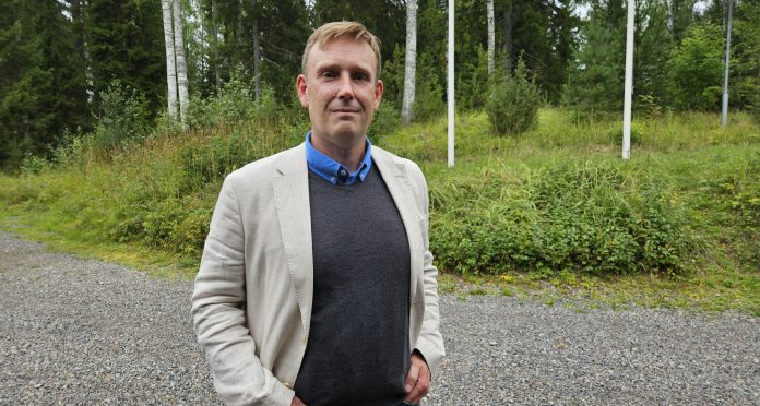 Antti Malinen by Emilia Karhu Kuvassa puolilähikuva miehestä jolla on harmaa villapaita ja valkoinen puvuntakki, taustalla näkyy puita ja nurmikkoa.