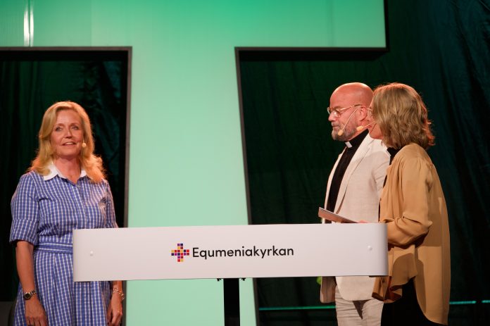 ElisabethThand Ringqvist by Kaj Aalto Kolme mikrofonia käyttävää henkilöä esiintyy lavalla vihreäksi valaistun taustan edessä. Etualan puhujakorokkeessa näkyvät värikäs ristisymboli ja teksti ”Equmeniakyrkan”.