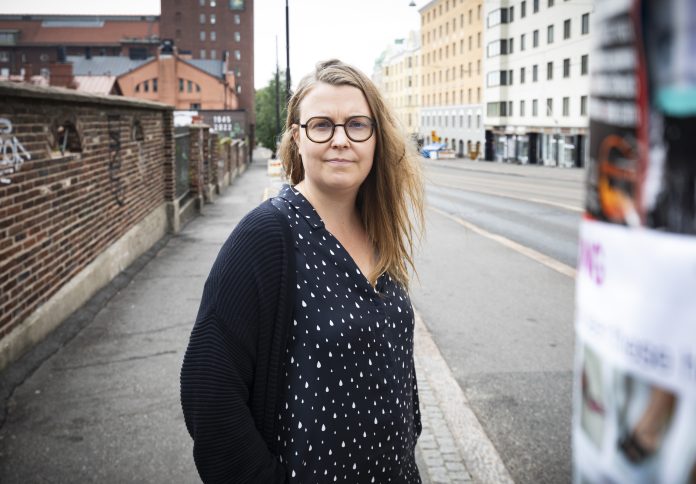 Sonja Finer Silmälasipäinen, pitkätukkainen henkilö seisoo kaupunkikadun reunalla ja katsoo kameraan. Taustalla näkyy tiiliseinä, eri-ikäisiä kerrostaloja ja lähes tyhjä katu.